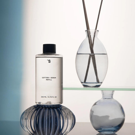 Sister's Aroma reed diffuser Cotton + Denim refill bottle with a glass diffuser vase on a reflective surface.