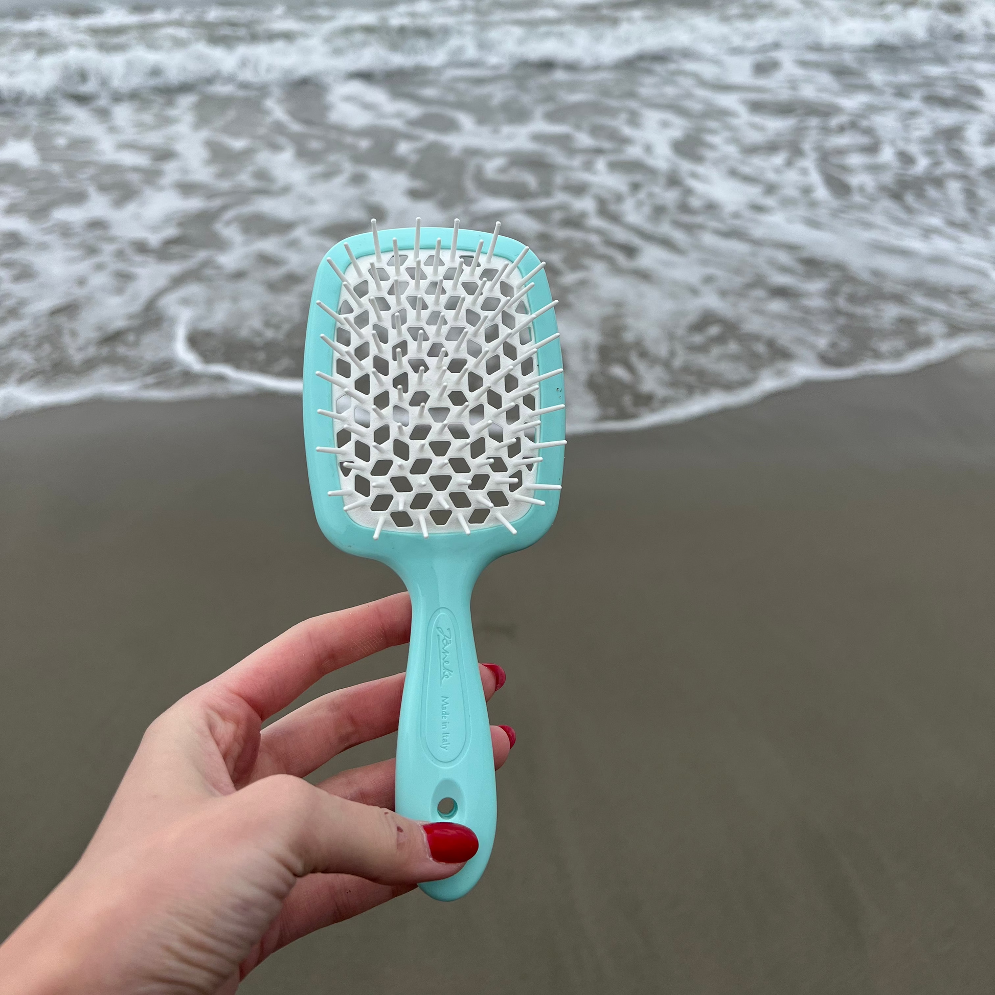 Hand holding an aqua blue Janeke hairbrush on a beach with ocean waves in the background