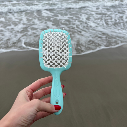 Hand holding an aqua blue Janeke hairbrush on a beach with ocean waves in the background